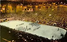 Far view of a ice hockey rink, with ice hockey players and a band standing at its centre. Spectators seated around the rink watch.