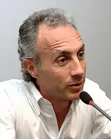 40-ish man with grey hairs, talking on a microphone.