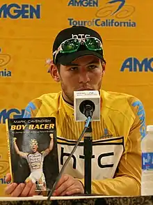 Cavendish sitting in front of a microphone holding a copy of his book Boy Racer