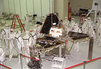 A group of scientists, all wearing white protective clothing, gather around a spacecraft as it is being folded into its launch position; a triangular pyramid shape.