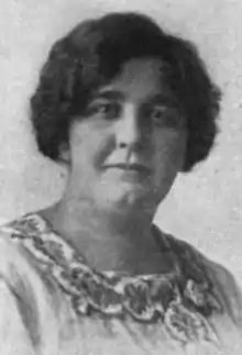 A black-and-white photograph of a white woman with dark wavy hair and a dimpled chin; she is wearing a dress or blouse with an embroidered square neckline