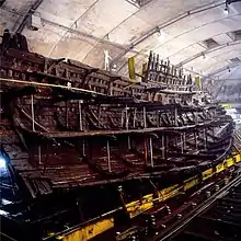 A diagonal section of a wooden ship resting upright on a yellow steel frame inside a building with bare, drab walls and sprinkler systems in the ceiling
