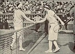 Lenglen and Wills shaking hands at the net while a person at the side holds their right hand up in a "stop" gesture