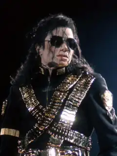 Jackson is wearing sun glasses and a black jacket with a golden metal rectangle on both sides of the collar, golden badges on both upper arms and a golden armband on his right arm under the badge. Two golden bandoliers cross his torso forming the letter "X". A third bandolier runs from his left shoulder to the bucket of his golden belt. He is frozen. His head is turned to the left. His hair is falling in his eyes and he is wearing a headset microphone. His skin is light