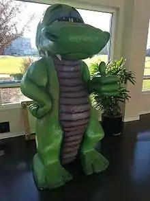 A photograph featuring the front right side of a statue of green lizard with a purple abdomen and chest. The lizard stands on two feet with its right arm on its hip and the left arm extended outward at the elbow, giving a thumbs up. His eyes are partially closed and several sharp teeth extend from the closed mouth.