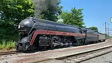 A black streamlined steam locomotive pulling a train with the trees and overhead lines in the background