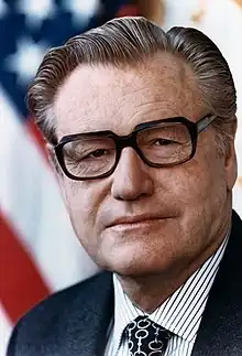 Posed head shot of a middle-aged man, dressed in a business suit. The American flag is visible behind him.