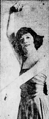 A white woman with short dark hair, in a dance pose, with one arm above her head and one across her front. She is wearing a sleeveless dress.