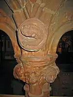 The top of a column and base of two arches. In the spandrel is a carving of an otter-like creature; the capital is decorated with a carved head and foliage