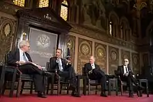 Four men in suits sit on chairs on a red stage in front of ornate gold and brown wall.