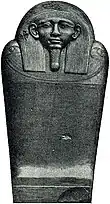 Black and white image of a dark stone coffin with a human face, the coffin stands upright facing the viewer.