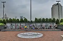 Ten large panels made of a dark stone are engraved with images from the Vietnam War. Flowers and wreaths lean against them. In front of the panels is a brick plaza with a liberty bell design.