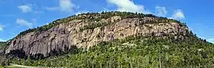 A low wooded mountain with exposed brownish stone cliffs, seen from across a road visible at the lower left. The base of the cliffs is higher at the right