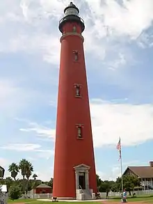A tall red lighthouse, seen from the point of view of someone on the ground, is seen framed by a blue sky, with palm trees and a flagstaff in the background.