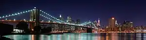 A brightly lit city at night. A suspension bridge lit up with bright blue lights crosses a still body of water in front of the city. The water reflects the blue light, causing rainbow-like stripes to appear on the surface where the lights of the bridge and the city meet.