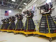Six rocket engines, consisting of a large bell-shaped nozzle with working parts mounted to the top, stored in a large warehouse with white walls decorated with flags. Each engine has several pieces of red protective equipment attached to it and is mounted on a yellow wheeled pallet-like structure.