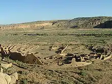 Daytime view looking down on a desert valley: in the near distance, a large semi-circular set of tumbled-down and ruined walls, greyish-yellowish brown in color. The far side of the ruins is a straight line, running left-right, roughly parallel to a line of cliffs in the far distance.