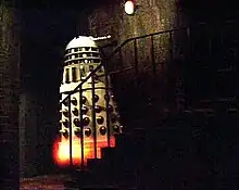 In a dark basement, a white Dalek (see previous description) appears to levitate up a small staircase of approximately seven stairs. The body of the Dalek is white, with shiny gold vertical slats and gold balls on its lower half. There is an orange-yellow glow at the Dalek's base.