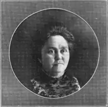 A white woman with hair in a bouffant updo, wearing a high-collared black dress with a lace yoke, in a circular frame