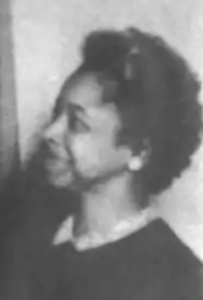 A middle-aged Black woman, smiling, wearing a dark top  with a light collar, and button earrings