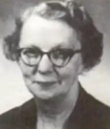 An older white woman wearing round glasses and a dark dress or jacket