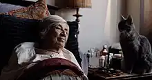 An old woman in bed with a cat standing on the bedside table