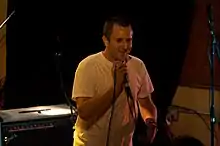 A man in a white T-shirt smiles and grasps a corded mic at its top