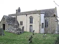 A small stone church seen from the southeast with a porch to the left, a protruding rendered south aisle in the middle, and a short chancel to the right. On the west gable is a bellcote, and in front of the church is a churchyard cross