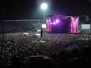 A stadium filled with spectators on the podiums and on the ground. In the middle is a stage with two giant pink 'L’ symbols flanking it. A large flood light is visible behind it.