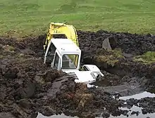 Photograph of a backhoe that is over fifty percent submerged in a large hole that it dug in a peat bog before falling in.