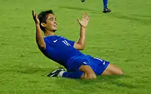Chhetri on his knees in a blue football shirt and blue football shorts. His arms are outstretched and he is smiling after just scoring a goal.