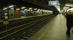 An indoor train station with two tracks running down the center. There are concrete platforms on both sides, with some people waiting.