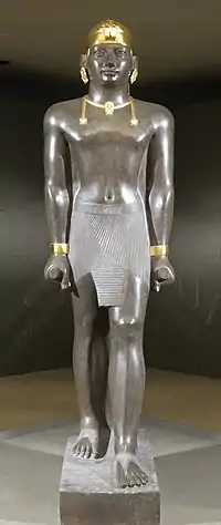Statue of Taharqa. His name appears on the center of his belt: 𓇿𓉔𓃭𓈎 (tꜣ-h-rw-q, "Taharqa"). The statue is 2.7 meters tall. Taharqa has a striding pose, the arms held tight, and holds the mekes staff. He wears a pleated kilt called shendjyt and on the head the double-uraeus skullcap, possibly signifying the double rule over Nubia and Egypt. (Louvre Museum, color reconstruction through pigment analysis).