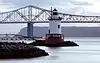 A red, white and black lighthouse at dusk with a large bridge in the background