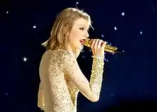 A young white woman in red lipstick and a bob hairstyle singing on a microphone, wearing a sparkling bodysuit