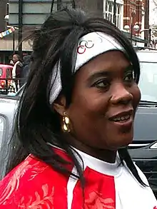 A brown-skinned woman with straight black hair, wearing a white headband and a gold hoop earring, looks off to one side