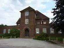 A photograph of a large derelict brick building from the 1930s. The front of the building has a large, rounded archway in the art deco style. The lower windows are boarded up, and the panes of many of the upper windows are smashed