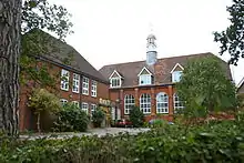 An L-shaped, red bricked building with a white bell tower. A tree is to the left of the photo.