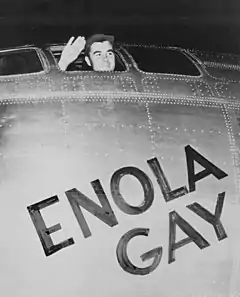 A man waves from the cockpit window of a plane with the words "Enola Gay" written on it