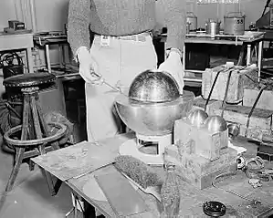 A re-creation of the Slotin incident. The inside hemisphere with the thumb-hole next to the hand is beryllium (replacing the uranium tamper in a Fat Man bomb), with an external larger metal sphere under it, of aluminium. The 3.5-inch-diameter (89&nbsp;mm) plutonium "demon core" (the same as in the Daghlian incident) was inside at the time of the accident, and would not be visible. However, its dimensions are comparable with the two small half-spheres shown resting nearby.