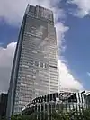 Ground-level view of a rectangular, glass high-rise; a smaller, circular building is in the foreground