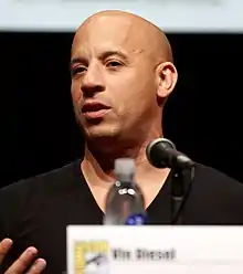 A bald, multi-racial man in a gray suit. Turned at a 45 degree angle from the camera, he squints in the direction of an off-camera light source, which illuminates his face.