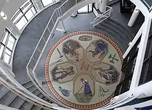 An aerial view of a mosaic depicting five people working, mining, farming -- all radiating out from the center like a flower. The are also tools of various trades like a hammer and a scythe surrounding the flower-like part.