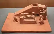 A Han-dynasty pottery model of two men operating a winnowing machine with a crank handle and a tilt hammer used to pound grain.
