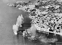 a black and white photograph of a ship at sea near a town; the ship is surrounded by smoke and spouts of water from explosions
