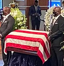 Two men wearing dark business suits stand on either side of a casket. The casket has been draped with a flag, a sign of military or government service in that country